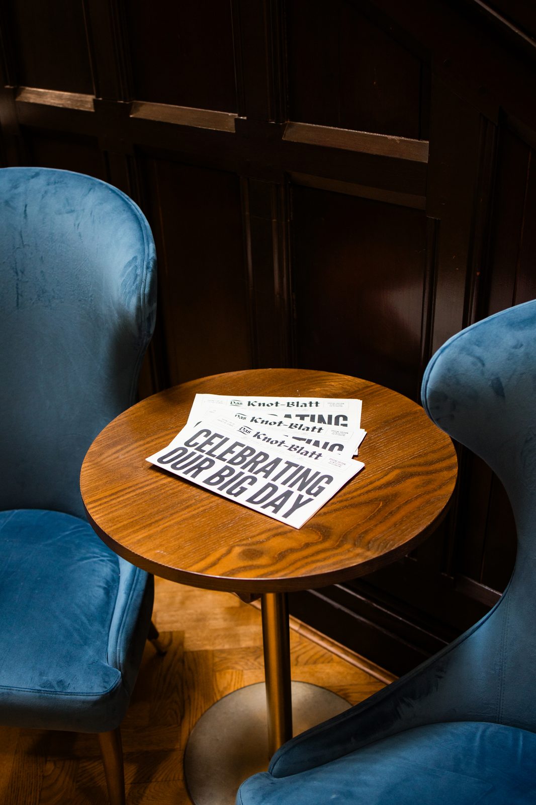 A stack of printed wedding newspapers on a small round table between two blue armchairs